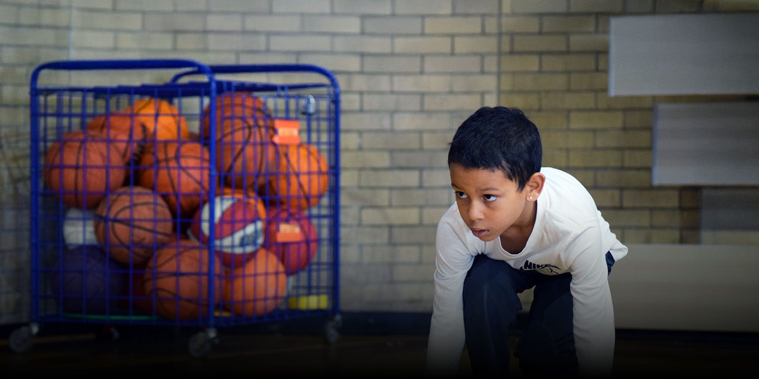 Student in a ready stance in the school gym, with a cart of basketballs in the background.