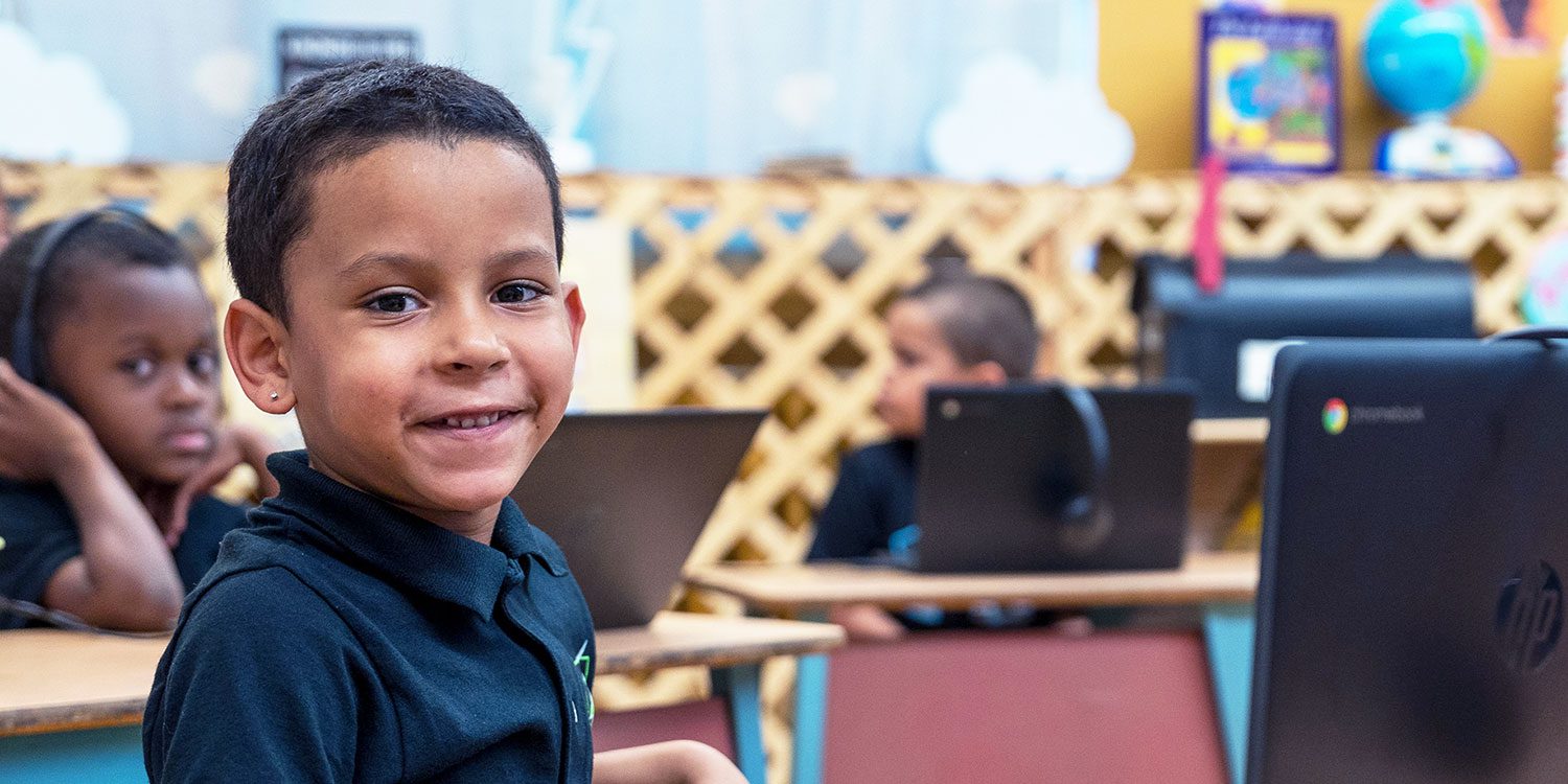 Smiling student at a desk in a classroom with laptops.